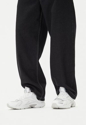 Bijele Reebok atletske tenisice nošene uz široke crne hlače, stojeći na običnoj bijeloj površini.