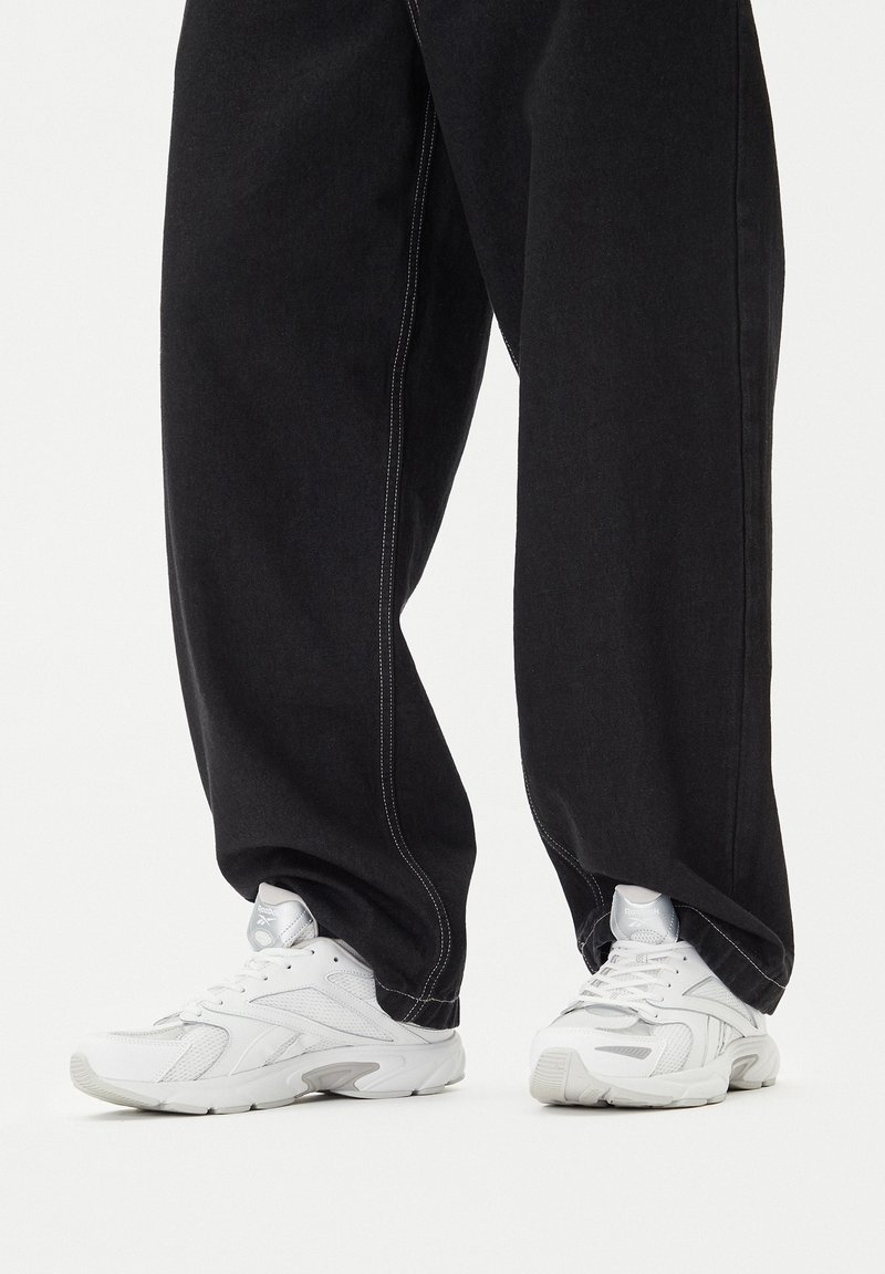 Baltos Reebok sportinės kedai dėvimi su laisvomis juodomis kelnėmis, stovint ant paprastos baltos spalvos paviršiaus.