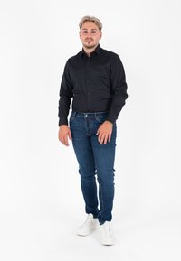 Camicia nera a maniche lunghe con bottoni, jeans elasticizzati blu scuro e sneakers bianche; modello in piedi contro uno sfondo bianco.
