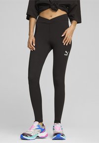 Leggings noirs avec une taille haute croisée et un logo Puma blanc. Tissu texturé et extensible. Portés avec des chaussures de sport multicolores.