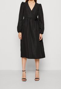 Svart omlottklänning med djup V-ringning, puffade långa ärmar och knytskärp i midjan. Slät textur och knälång design. Bärs med klackar med ankelsnören.