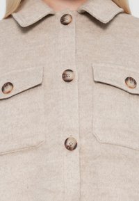 Bež villasegu shirt-jakett, millel on krae, kahe rinna-tasku ja suurte, ümmarguste, kontrastsete nööbidega. Pehme tekstuur harjatud viimistlusega.
