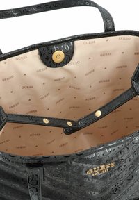 Sac fourre-tout noir avec motif en relief, accents dorés, fermeture à pression et intérieur beige comportant le logo "GUESS" répété et des motifs triangulaires.