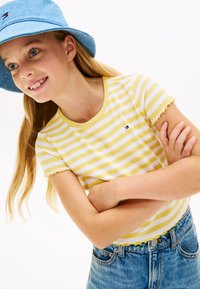 Κίτρινο και λευκό ριγέ T-shirt για νέους με διπλωμένα μανίκια, συνδυασμένο με ψηλόμεσο μπλε τζιν σορτς. Διακριτικό μπλε καπέλο κουβάς.