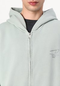 Felpa con zip di colore verde chiaro, dotata di cappuccio, texture a costine e dettaglio logo sul lato sinistro del petto. Presenta una zip argentata e cuciture lungo le cuciture.