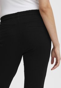 Černé kalhoty s elastickým pasem a hladkým povrchem, se dvěma malými zadními kapsami a přiléhavým střihem nohavic.