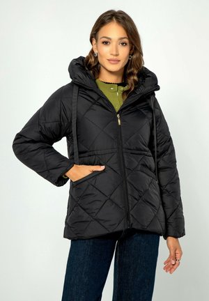 Zwarte gewatteerde pufferjack met een capuchon, met een voorrits, trekkoorden en zijzakken. Draag het over een groene top en donkere jeans.
