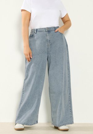 Person trägt hellblaue Jeans mit weitem Bein und weiße Turnschuhe, eine Hand in der Tasche, vor schlichtem weißem Hintergrund.