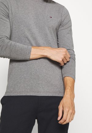 Mand justerer ærme på tætsiddende grå langærmet skjorte med lille broderet logo, iført sorte bukser, foran ensfarvet baggrund.