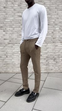 Persoană îmbrăcată cu o cămașă albă cu mâneci lungi, pantaloni bej cu șnur și mocasini negri cu ciucuri, stând lângă un zid de cărămidă deschisă.