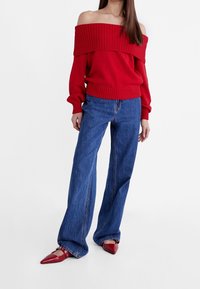Femme portant un pull rouge à épaules dénudées, un jean large bleu et des ballerines rouges à bouts pointus, debout devant un fond blanc uni.
