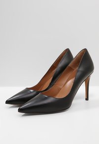 Černé kožené lodičky na vysokém podpatku s špičatou špičkou, hladkou texturou a štíhlým jehlovým podpatkem, se světlým bežovým stélkou.