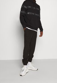 Svart hoodie med logoband, ficka fram och dragsko i huvan; matchas med mörka joggers och vita sportskor. Enkel, avslappnad design.