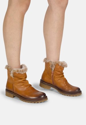 Piernas con botines de cuero marrón con ribete de piel y cremalleras laterales sobre un fondo claro.