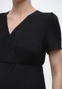 Κοντινό πλάνο ενός ατόμου που φοράει μαύρο φόρεμα με κοντά φουσκωτά μανίκια και V-λαιμόκοψη, εμφανίζοντας τον άνω κορμό και το χέρι.