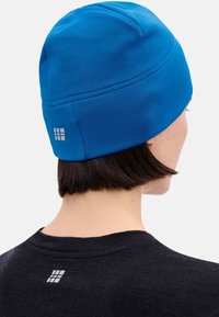 Persoon met kort donker haar, een felblauwe muts en een zwart shirt, beide voorzien van een klein wit rasterlogo op de achterkant.