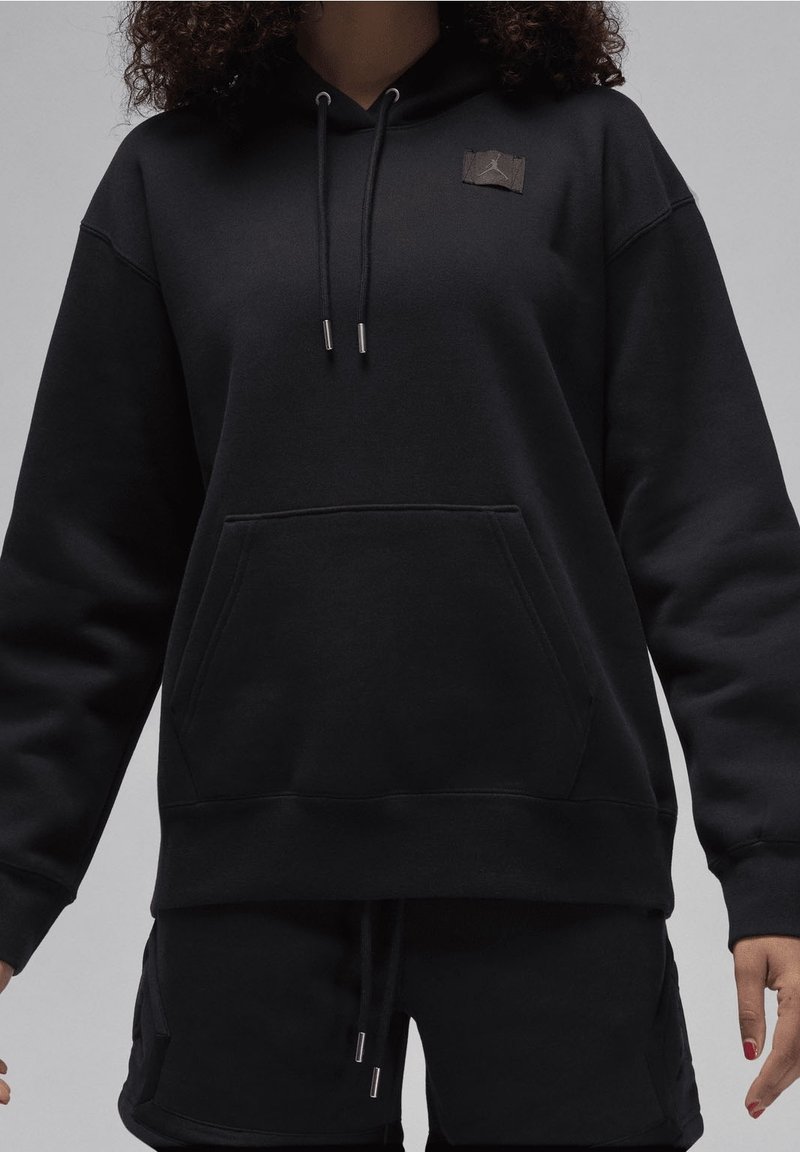 Personne portant un sweat à capuche noir avec une poche avant et des cordons, associé à un short noir, debout devant un fond uni.