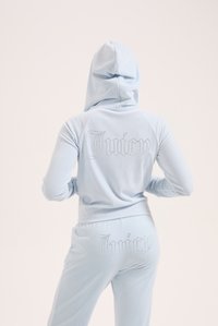 Svetlomodrý zamatový teplákový komplet s kapucňou. Logo "Juicy" zdobené krištáľmi sa nachádza na zadnej strane mikiny a nohavíc. Hladký povrch.