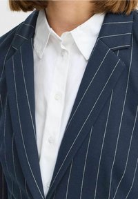 Granatowa marynarka w pinstripe z białymi paskami i klapami z wycięciem, zestawiona z gładką białą koszulą na guziki. Gładka struktura tkaniny.