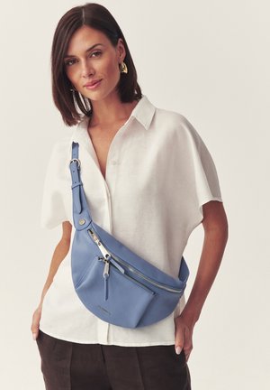 Lühikeste tume juustega naine kannab valget särki ja sinist nahast ristseljakotti, poosib ühevärvilise tausta ees.