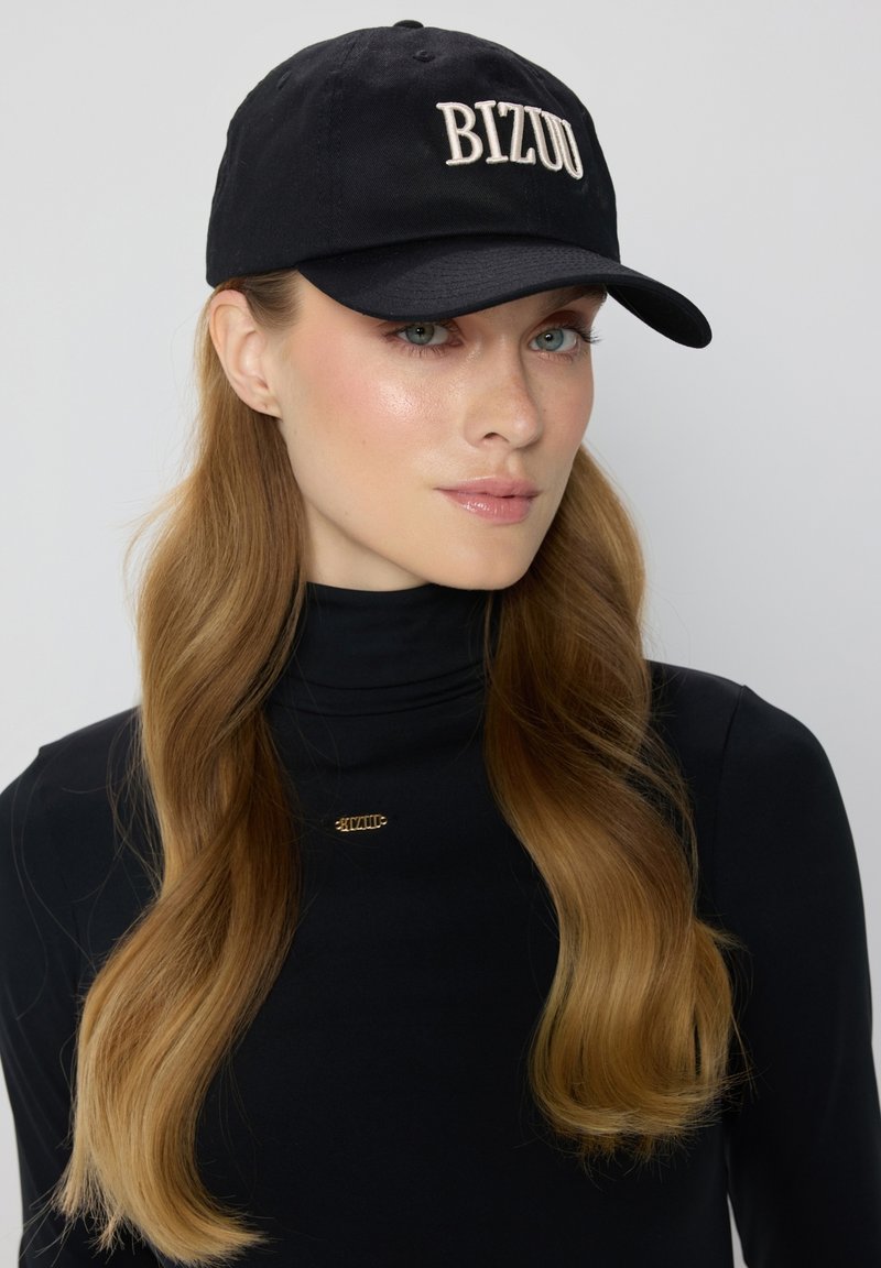 Zwarte baseballpet met "BIZUU" geborduurd in lichtbeige aan de voorkant. Model draagt een zwarte coltrui en heeft lang, golvend haar.