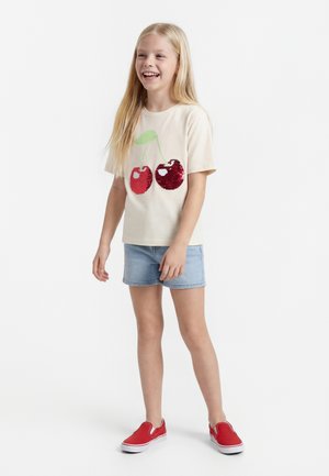 Young girl smiling, wearing beige t-shirt with two sequined cherries, light denim shorts, and red slip-on shoes, standing on white background.