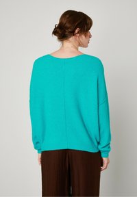 Turquoise oversized sweater met een ruime ronde rug, lange mouwen en een geribbeld zoom, gecombineerd met bruine plooibroeken. Zachte textuur.