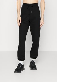 Pantalones de chándal negros con cintura elástica ajustable, piernas ajustadas, puños acanalados y una marca mínima, combinados con zapatillas de deporte negras y calcetines blancos.
