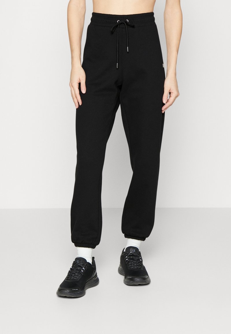Pantalones de chándal negros con cintura elástica ajustable, piernas ajustadas, puños acanalados y una marca mínima, combinados con zapatillas de deporte negras y calcetines blancos.