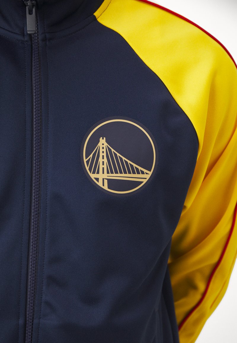 Felpa da tuta blu navy con maniche gialle, caratterizzata da un logo rotondo dorato di un ponte sul lato sinistro del petto. Chiusura con zip e texture morbida.