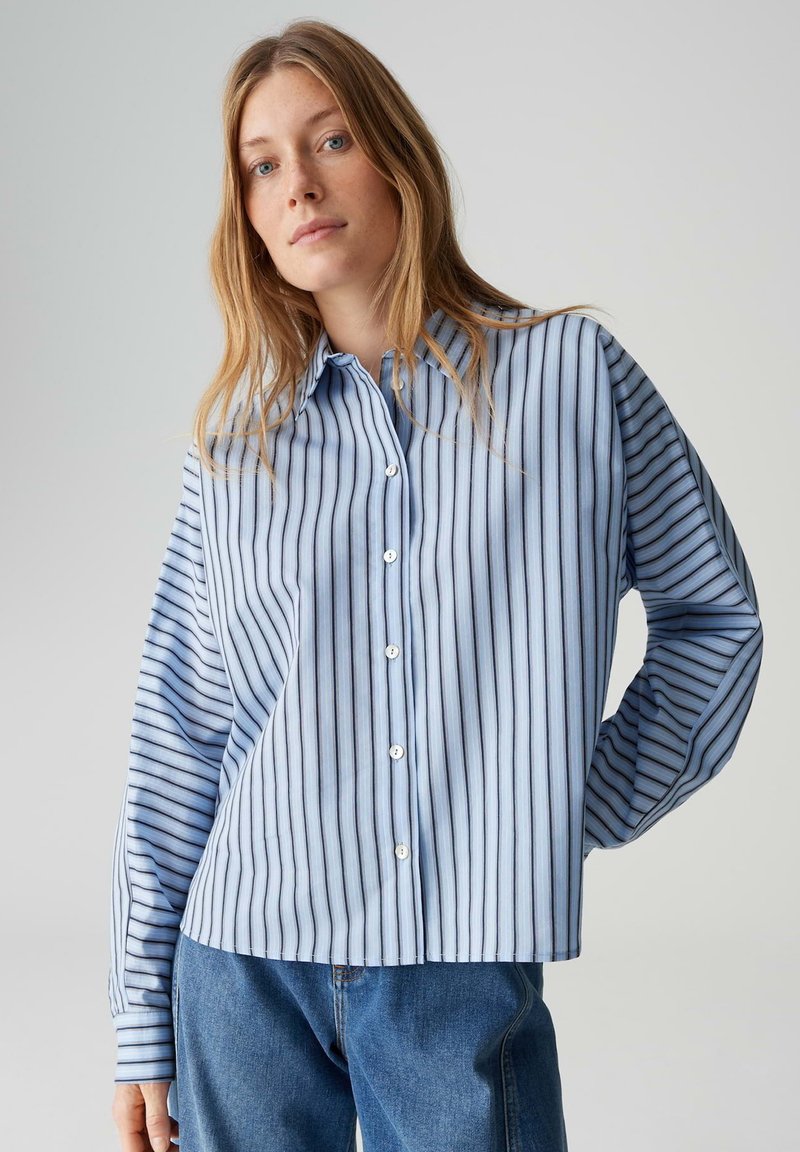 Chemise à manches longues bleu clair avec des rayures blanches horizontales, boutonnée sur le devant, et coupe décontractée. Portée avec un jean en denim.