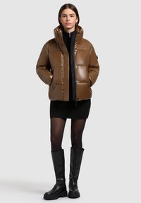 Jachetă puffer maro cu guler înalt, textură lucioasă, nasturi tip capcană și două buzunare frontale, purtată peste o rochie neagră cu dungi și cizme până la genunchi.