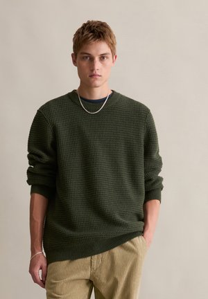 Groene gebreide trui met een gestructureerd patroon, ronde hals en ribgebreide manchetten, gecombineerd met beige corduroy broek. Zilveren kettingaccessoire zichtbaar.