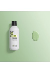 Weiße Plastik-Shampooflasche mit einem schwarzen Deckel, beschriftet mit "KMS Add Volume", neben einem mintgrünen Klecks Shampoo auf einem hellgrünen Hintergrund.