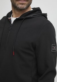 Crna zip-up majica s kapuljačom, prednjim džepom i crvenim detaljima na šniri. Na lijevoj rukavu ima zakrpu s detaljima logotipa.