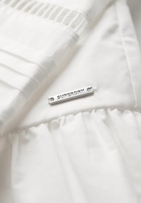 Biały materiał z plisowanymi detalami i falbaną. Przymocowana metalowa etykieta "SUPERDRY" w kolorze srebrnym. Miękka tekstura, czysty design.