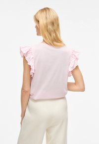 Helles rosa T-Shirt mit Rüschensäulen, aus Baumwollmaterial, lockere Passform. Sichtbare Nahtdetails und runder Ausschnitt. Getragen mit cremefarbenen Hosen.