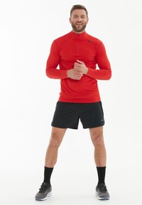 Rød langærmet træningsshirt med høj krave, parret med sorte shorts. Model iført sorte sokker og grå sneakers.