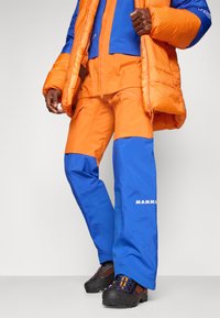 Ανδρας που φοράει πορτοκαλί και μπλε παντελόνι εξωτερικών δραστηριοτήτων, μαύρα παπούτσια πεζοπορίας και πορτοκαλί μπουφάν, στέκεται μπροστά σε ουδέτερο φόντο.
