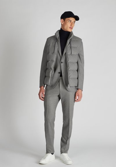 Giacca a vento grigia sopra un completo grigio, con colletto alto e cerniera. Abbinata a sneakers bianche, l'outfit presenta un design elegante.