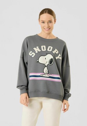 Junge Frau trägt grauen Sweatshirt mit Snoopy-Cartoon und weißen, rosa und blauen Streifen, kombiniert mit hellen Hosen, lächelt in die Kamera.