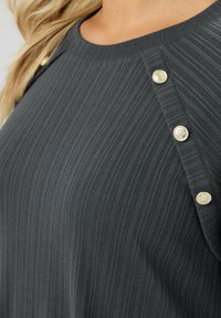 Primo piano di un top in tessuto a coste scuro con bottoni decorativi dorati lungo la cucitura della spalla e parte dei capelli biondi visibili.