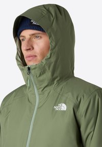 Oliivinvihreä huppari, jossa on vetoketju edessä, rint Logos ja teksturoitu kangas, osittain avattu ja tiukasti istuva huppu.