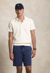 Polo skjorte i off-white med korte ermer, liten logo; blå snøring shorts; mørkeblå caps; uformell klokke på handleden. Nøytral bakgrunn.