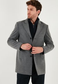 Cappotto grigio in lana con colletto a punte, due tasche frontali e chiusura con bottoni. Sotto, una giacca blu con zip è parzialmente visibile.
