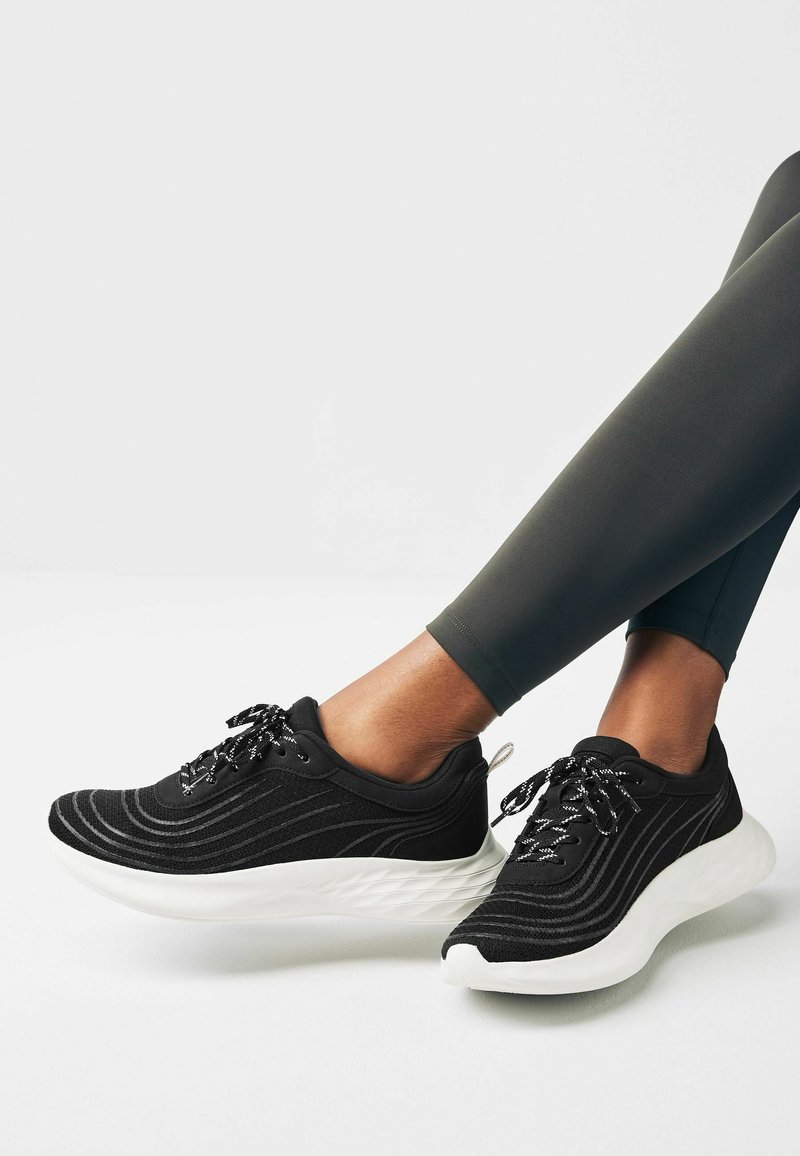 Black athletic shoes with a textured upper, white sole, and reflective laces. Features a wave pattern on the fabric for added detail.