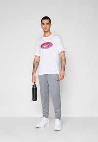 Hvit T-skjorte med en rød og blå gradient Nike-logo, grå joggebukser, hvite joggesko, holder en svart Nike vannflaske.