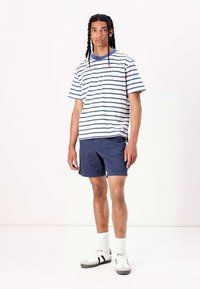 Stribet bomulds t-shirt i hvid og marineblå, parret med marineblå shorts. Har korte ærmer, rund hals og afslappet pasform. Bæret med hvide sokker og sneakers.