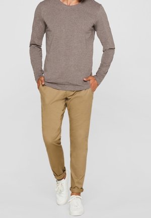 Homme portant un pull brun à manches longues, un pantalon kaki retroussé et des baskets blanches, debout les mains dans les poches devant un fond uni.