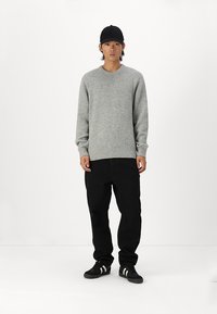 Pelēks adīts džemperis ar apaļu kakla izgriezumu, ribotām piedurknēm un apakšmalu, kombinēts ar melniem džinsiem un melniem skriešanas apaviem ar baltām svītrām.